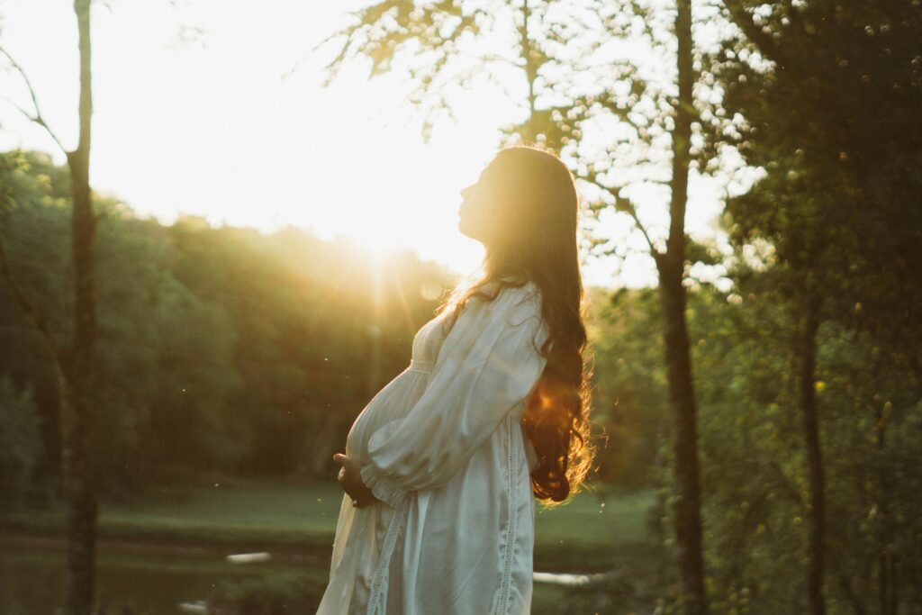 pexels-photo-19738355-19738355 Pregnant woman in white dress enjoying a sunset in a serene forest setting.