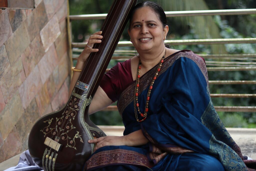 pexels-photo-5804658-5804658 Smiling woman in traditional Indian attire holding a tanpura outdoors.